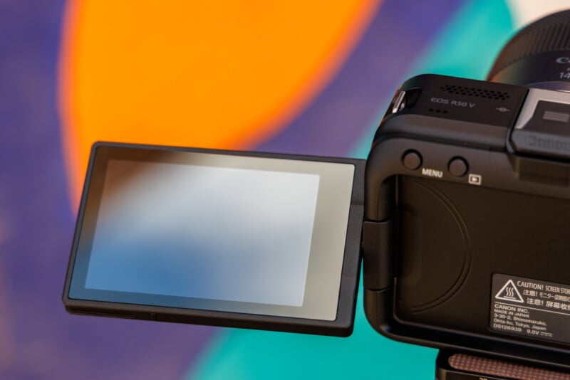 Close-up of an open camera with a flipped-out LCD screen. The background is colorful, featuring vivid orange and purple shapes.