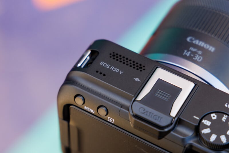 Close-up of a Canon EOS R50 V digital camera. Visible features include a textured top surface, a screen hinge, a mode dial, and a lens with "14-30" marking. The background is blurred with soft colors.