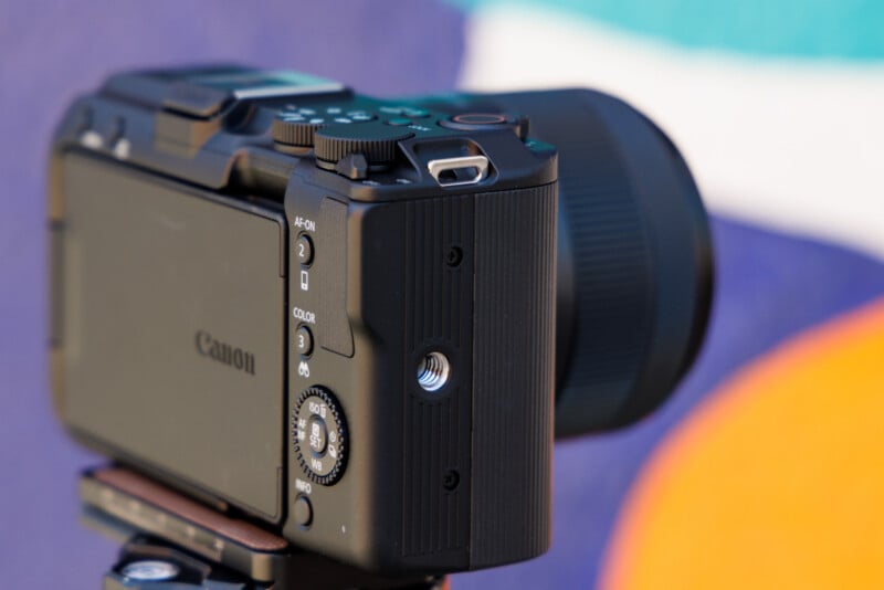 Close-up of a black Canon camera mounted on a tripod, focusing on the side buttons and lens. The background features blurred, colorful abstract shapes in blue, white, and orange.
