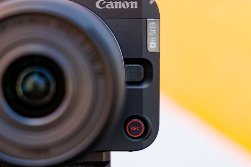 Close-up of a Canon EOS R camera with focus on the "REC" button, located near the bottom right. The lens is prominently visible on the left side. The background is blurred with shades of yellow and white.