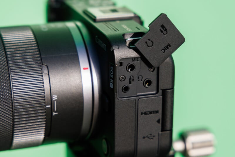 Close-up of a digital camera showing various input ports, including microphone, HDMI, and USB. The camera is black with a lens on the left side, and a small flap is open, revealing the ports. The background is a solid light green.
