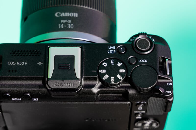 Close-up view of a Canon EOS R50 V camera on a turquoise background, showing the top control dials and buttons. The lens attached is an RF-S 14-30mm.