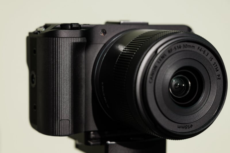 Close-up of a black digital camera with a zoom lens attached, positioned on a tripod. The camera's texture and lens details are visible, set against a muted green background.
