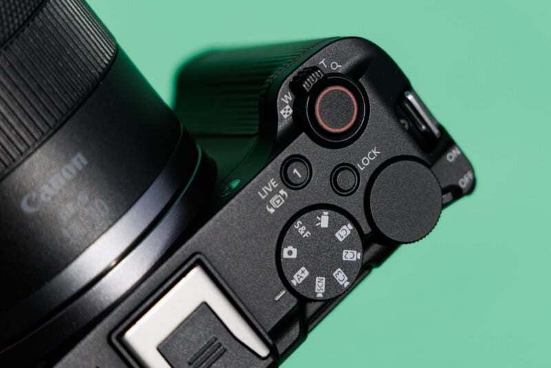 Close-up of a digital camera with its control dials and buttons visible. The camera is angled on a green background, highlighting the textured buttons and adjustment knobs around the shutter release button.