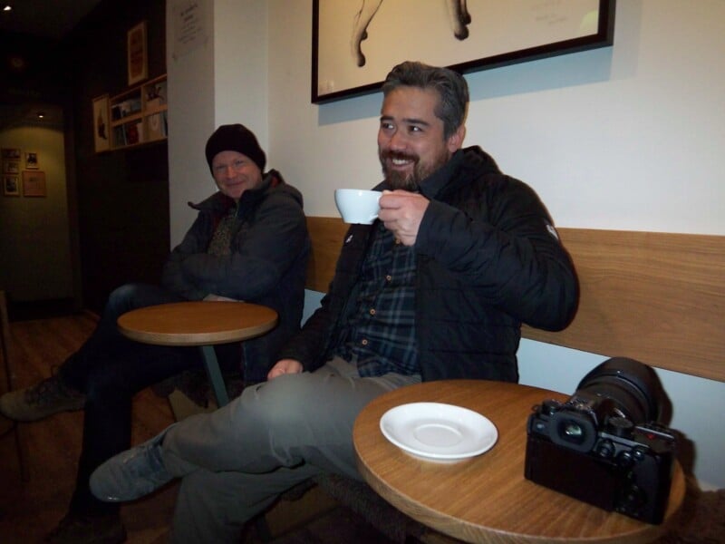 Two people sit at a café. One smiles while holding a cup of coffee, with a camera on the table. The other person wears a beanie and jacket, sitting relaxed. Tables and framed pictures are visible in the background.
