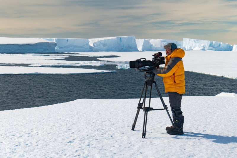 A person in a yellow jacket films a snowy, icy landscape with a professional video camera on a tripod near dark water and large icebergs under a cloudy sky.