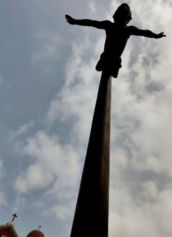 A tall statue of a person with outstretched arms stands atop a pole against a cloudy sky. A building with a cross is visible in the lower left corner of the image.