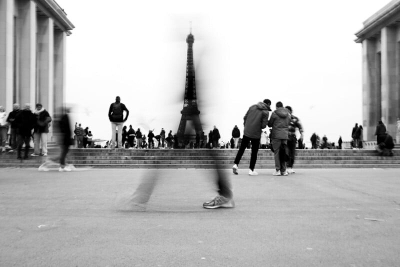 A black and white photo of people walking and gathering near the Eiffel Tower in Paris. The foreground shows a blurred figure in motion, with others standing or talking in the background.