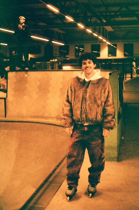 A person rollerblading indoors, wearing a furry jacket and standing near a skate ramp. Another person is visible in the background on the ramp. The setting is warmly lit, with an industrial ceiling and lights above.