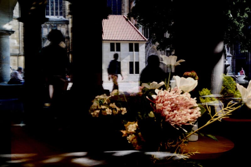 A bouquet of flowers in the foreground with silhouettes of people and an outdoor scene, including a small building and columns, visible through a window with reflections.