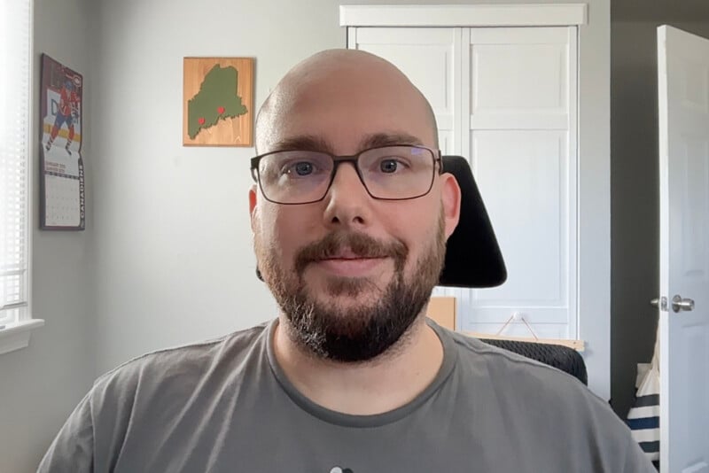 A bald man with glasses and a beard smiles at the camera. He is wearing a gray shirt and sitting in an office chair. Behind him are a closet, an open white door, and items on the wall, including a map and artwork. Light filters in through a window.