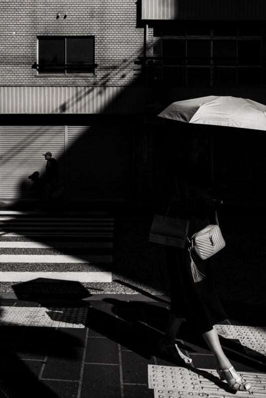 A person holding an umbrella and a handbag walks through a crosswalk in deep shadow, while another person walks in the background along a sunlit brick building. The image is black and white with strong contrasts.