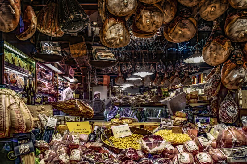 A bustling Italian market stall filled with hanging cured meats, cheeses, and fresh pasta. Two people work behind the counter, surrounded by shelves of delicacies and a vibrant display of food products.
