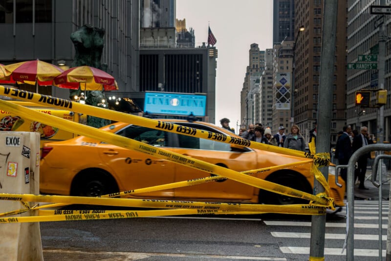 A yellow taxi cab speeds through a New York City street intersection, partially blocked by yellow caution tape, with pedestrians and tall buildings in the background.
