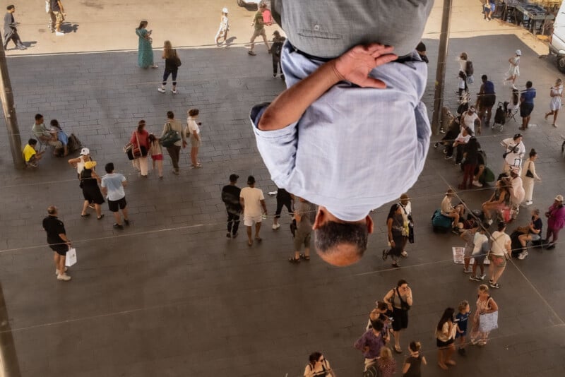 A man in a light blue shirt is reflected upside-down in a large mirror above a busy plaza where people walk, gather, and sit, creating an interesting visual illusion.