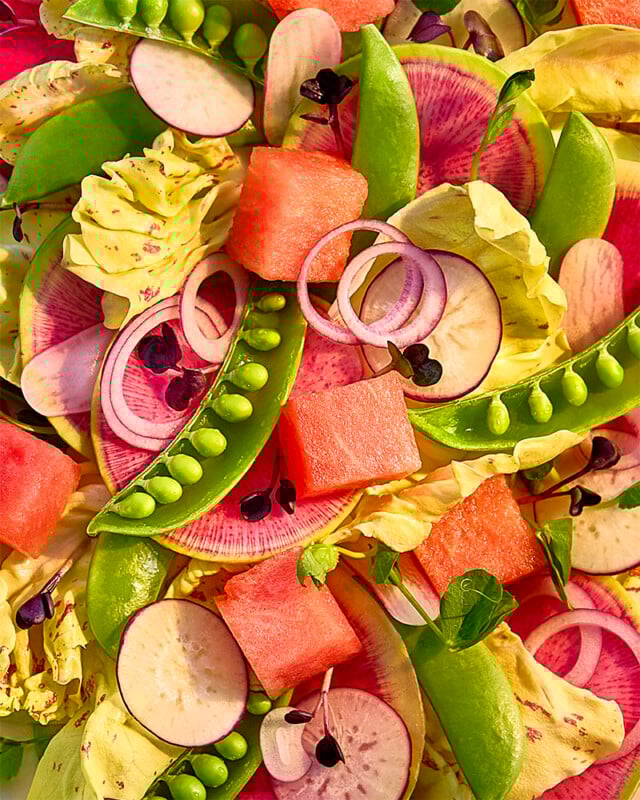 A colorful salad with lettuce, watermelon cubes, sliced radishes, snap peas, red onion rings, watermelon radish slices, and microgreens arranged closely together.
