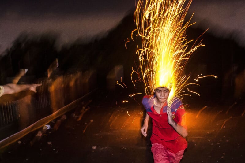 A boy in a red shirt runs at night with bright yellow fireworks shooting up from his head, creating a dramatic trail of sparks against a dark, blurred background.