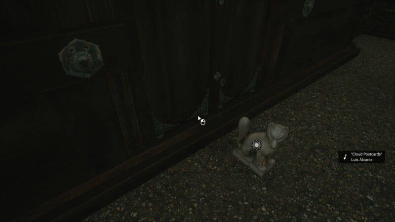 A small, cat-shaped stone statue is placed on a pebble-textured surface next to a dark wooden cabinet. The background is dimly lit, highlighting the contrasting textures of the materials.