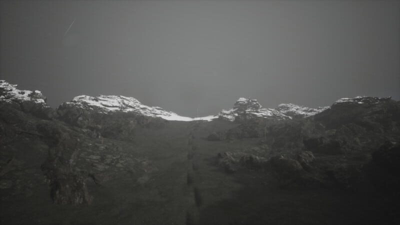 A rugged mountain landscape under an overcast sky. The rocky terrain is dusted with patches of snow and features a narrow, worn path leading upward through the center.