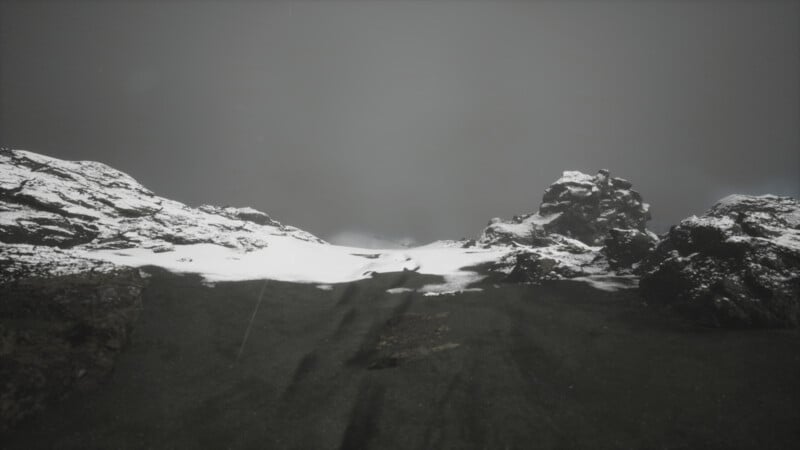 A desolate, snow-covered landscape with rugged rocks and a dark, overcast sky. Snow patches rest unevenly on the ground, creating a stark contrast against the rocky surface.