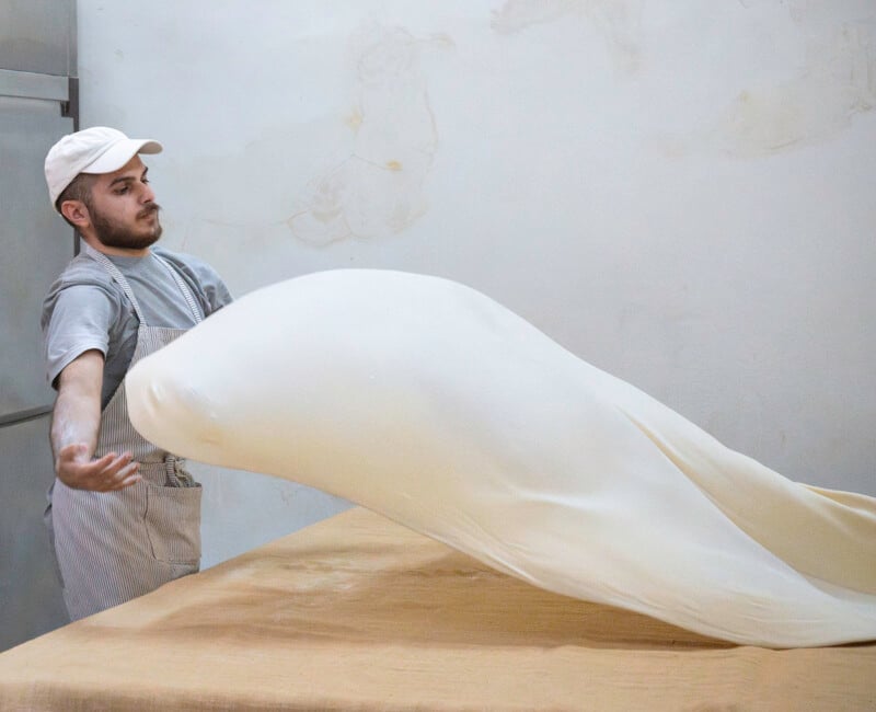 A man wearing a cap and apron stretches and tosses a large sheet of dough over a table in a kitchen with plain walls.