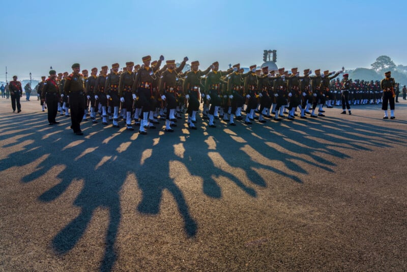 A large group of uniformed soldiers marches in formation on a sunlit parade ground, casting long shadows on the pavement. The sky is clear, and a few trees and buildings are visible in the background.