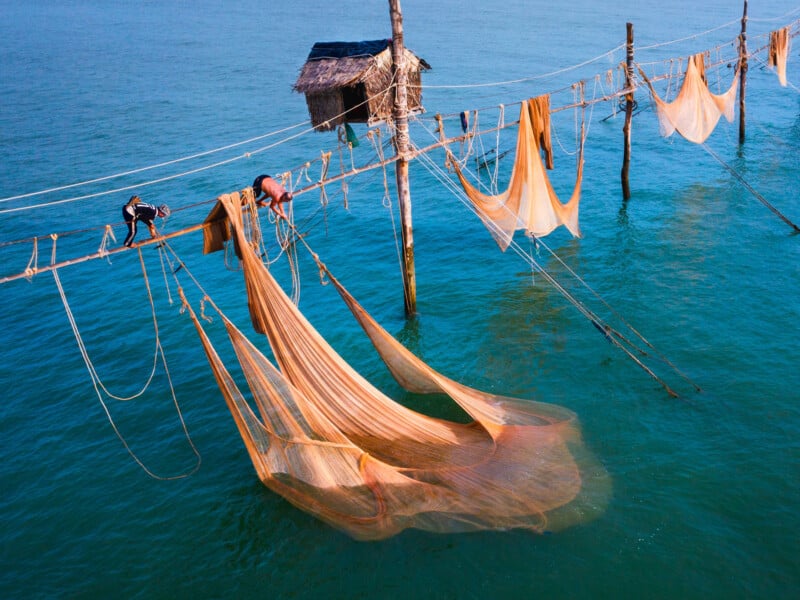 Two people work with large, orange fishing nets suspended over turquoise water, attached to wooden poles with a small hut on stilts in the background.