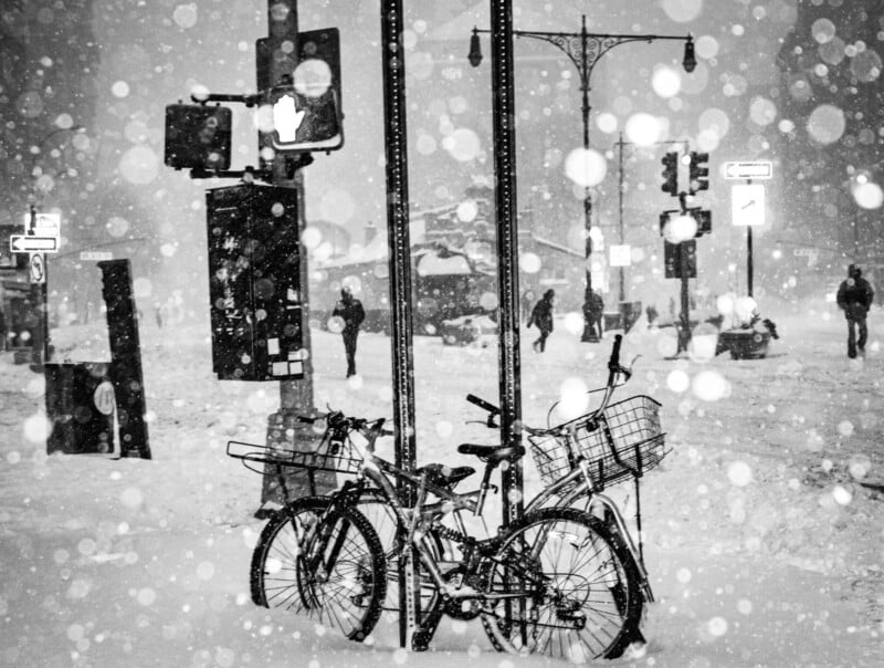 Two bicycles with baskets are locked to a street pole during heavy snowfall on a city street. Pedestrians walk in the background amid blurred traffic lights and street signs. Snowflakes fill the scene.
