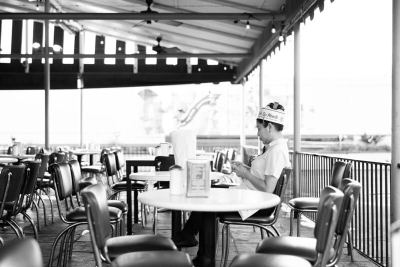 A person wearing a hat and uniform sits alone at an outdoor diner table, surrounded by empty chairs under a covered patio. The scene is in black and white.
