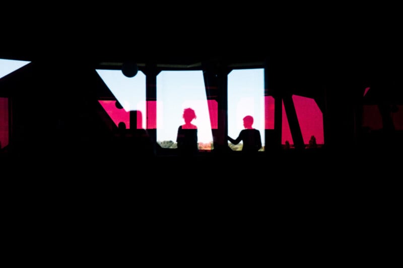 Two silhouetted figures stand by large windows with red and blue panels, looking outside. The figures are in shadow, and daylight streams through the windows, creating a strong contrast.