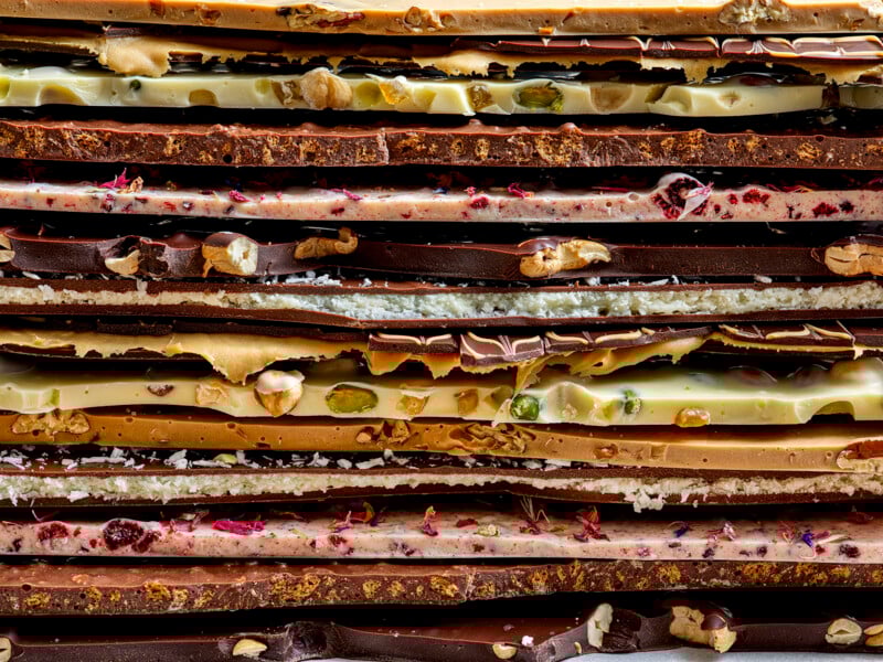 A close-up view of stacked chocolate bark pieces in various flavors, including dark, milk, and white chocolate, with assorted nuts and dried fruit visibly embedded in the layers.