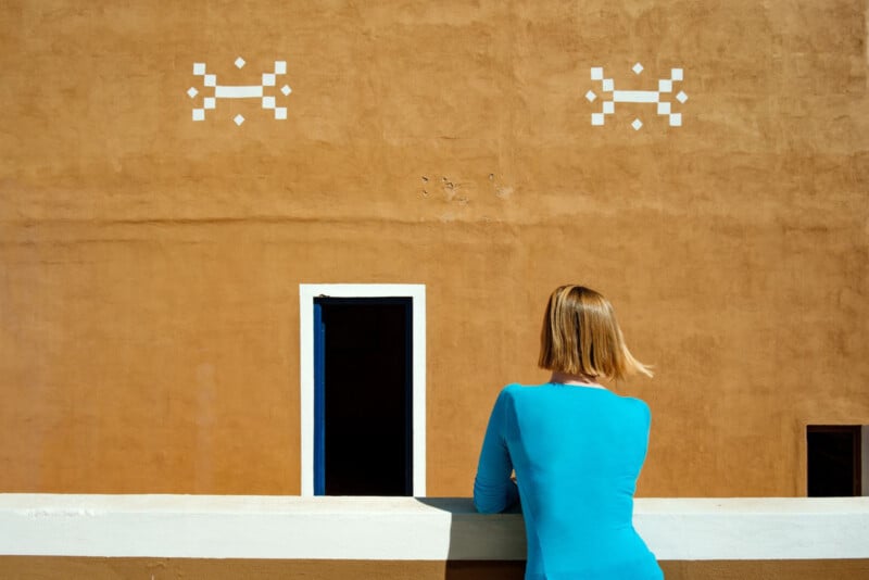 A woman with short blonde hair, wearing a blue shirt, leans on a cream-colored ledge, facing a mustard-yellow wall with a black open doorway and two white geometric designs near the top.