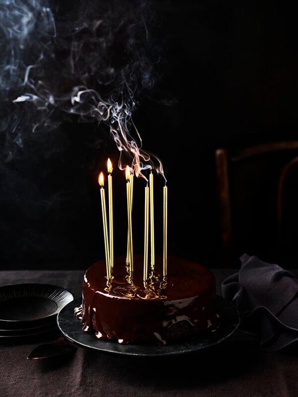 A chocolate cake with a glossy glaze sits on a dark plate. Five tall, gold candles burn on top, with trails of smoke rising. The background is dark, with empty plates and a napkin beside the cake.