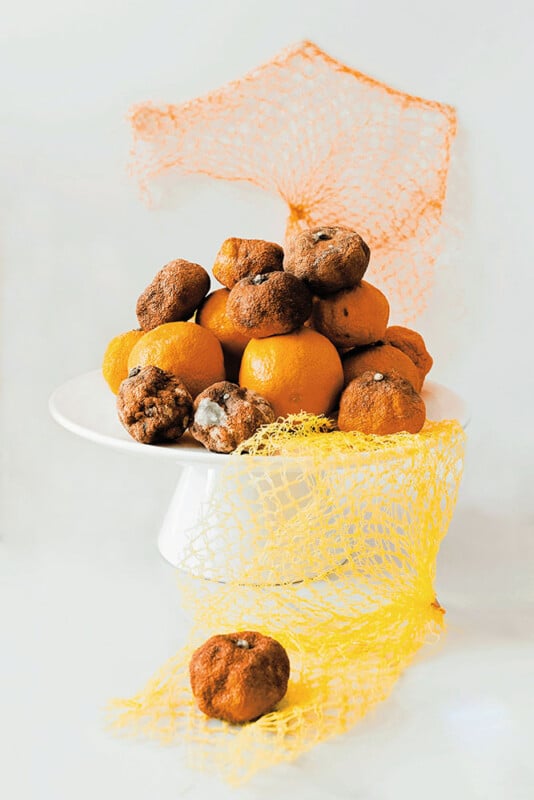 A white cake stand holds a pile of fresh and moldy oranges, surrounded by yellow and orange plastic netting, against a light background.
