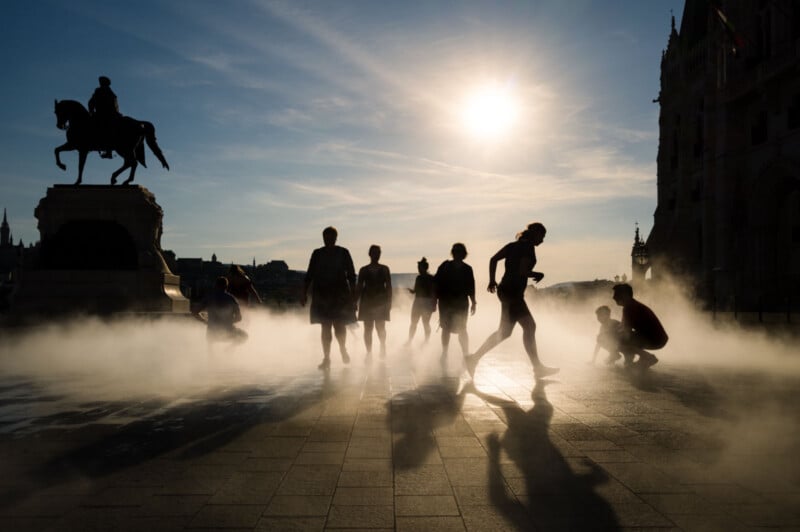 Silhouettes of people standing and crouching in mist on a plaza at sunset, with a statue of a rider on horseback and historic buildings in the background. Long shadows stretch across the ground.