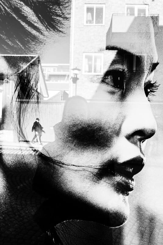 Black and white double exposure image of a woman's face in profile blended with an outdoor urban scene where a person walks near buildings under sunlight.