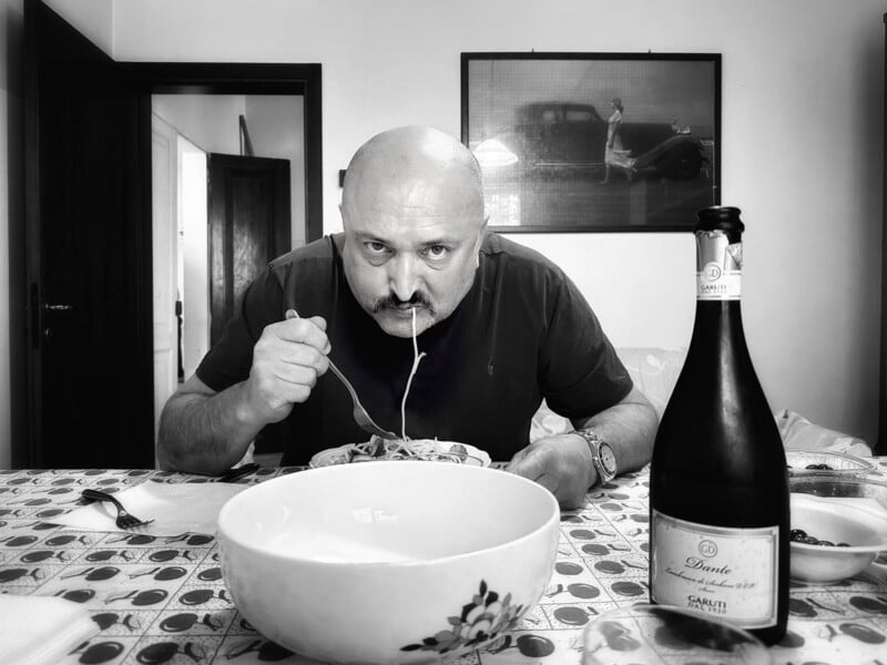 A bald man sits at a table eating spaghetti, staring intently at the camera. A large bowl and a bottle of sparkling wine are on the table. The black-and-white photo shows a vintage car picture on the wall behind him.