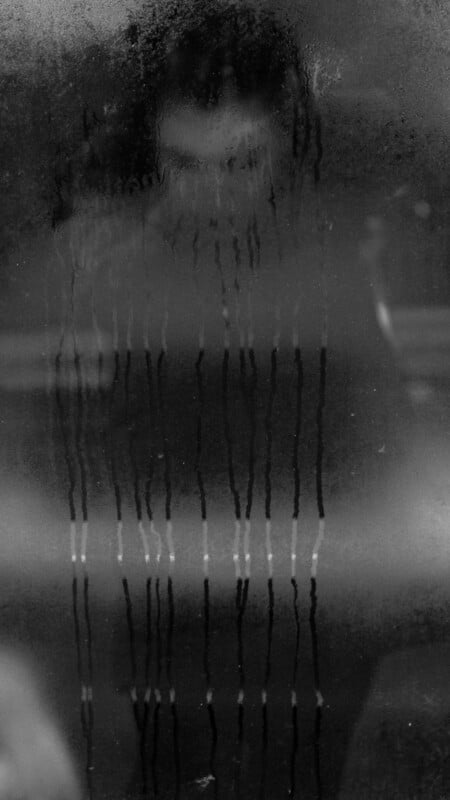 A shadowy figure is visible through a fogged glass pane with water streaks running down it. The person's features are blurred and obscured by condensation and darkness.