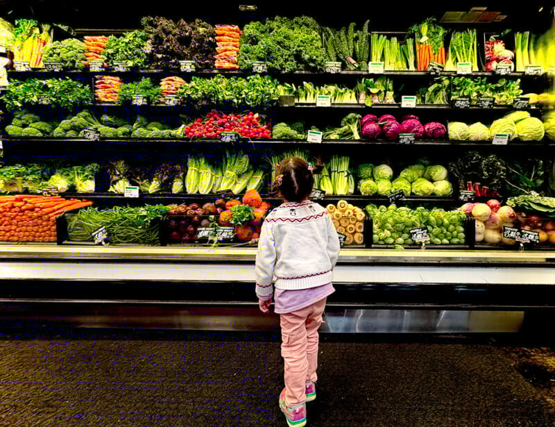 A young child in colorful clothes stands facing shelves full of fresh vegetables at a grocery store, with leafy greens, carrots, cabbages, and other produce neatly arranged in rows.