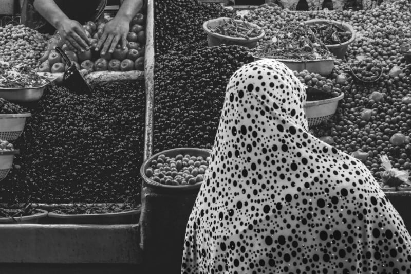 A person wearing a polka dot patterned headscarf faces a market stall filled with various fruits and vegetables, while another person organizes produce with their hands. The image is in black and white.