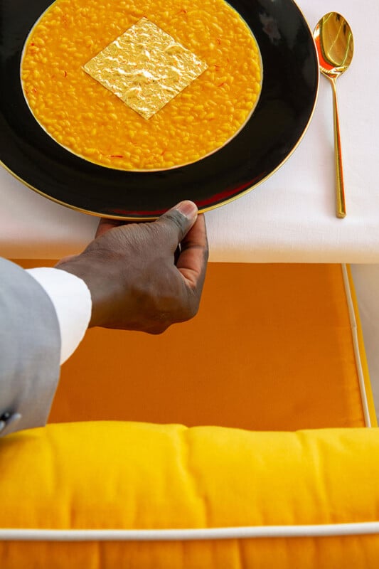 A person in a suit places a black plate with saffron risotto topped with a square of gold leaf on a white tablecloth, beside a gold spoon and a yellow cushioned seat.
