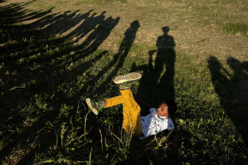 A child in yellow pants lies on the grass with one leg raised, while long shadows of several people stretch across the ground in the sunlight.