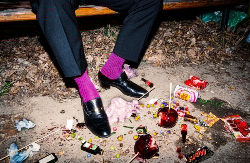 A person wearing black dress shoes and bright purple socks sits on a bench surrounded by scattered candies, lollipops, broken candy apples, and snack wrappers on the ground covered with dry leaves.