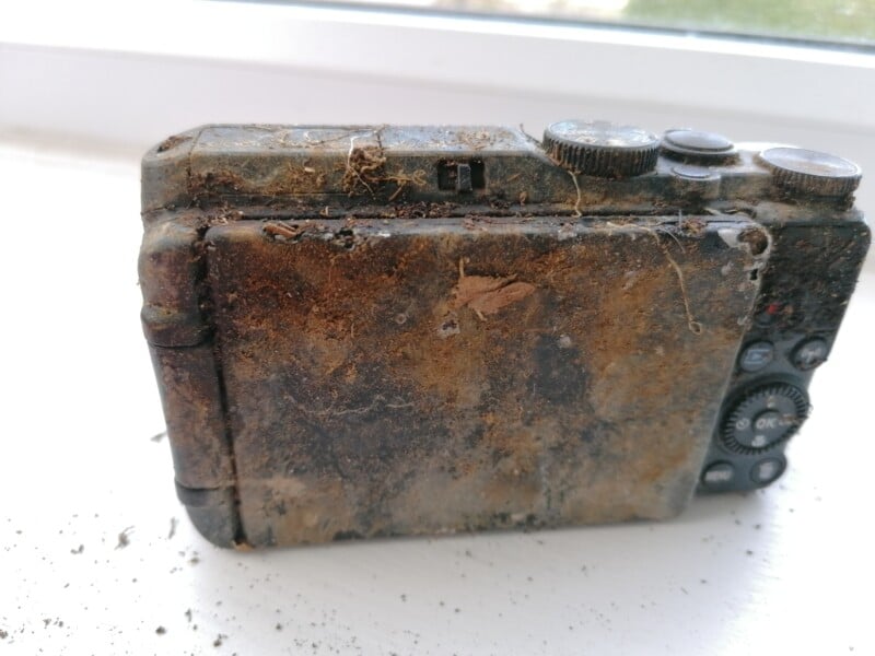 A heavily rusted and dirt-covered digital camera sits on a white surface, showing significant signs of damage and neglect. The buttons and dials are encrusted with grime and the screen is barely visible.