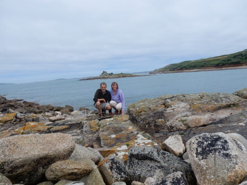 A man and a woman crouch together on rocky terrain by the sea, with an island and green hills visible in the background under a cloudy sky.