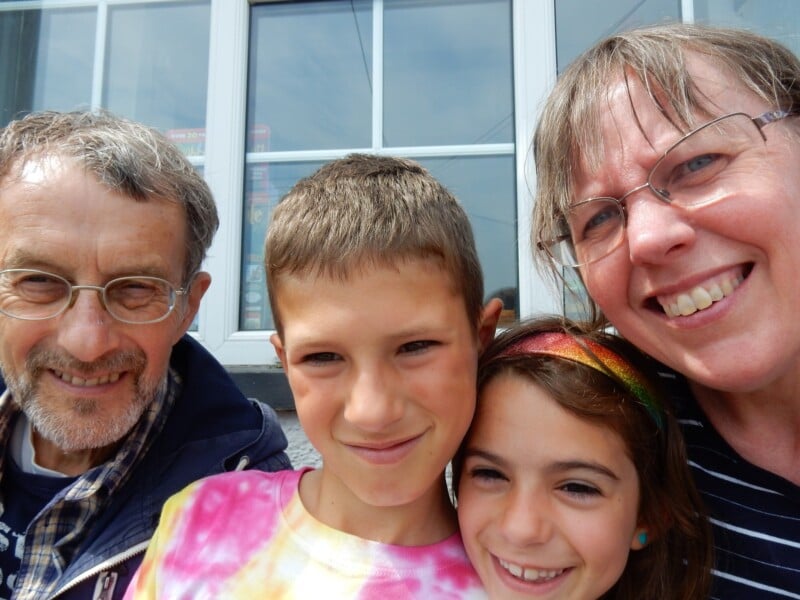 Four people, two adults and two children, smiling and posing closely together for a selfie in front of a white-framed window. The adults wear glasses, and one child wears a rainbow headband.