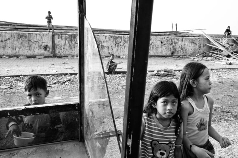 Three children stand by a glass window, two girls facing forward and a boy looking through the glass, holding a bowl. In the background, people are near a wall surrounded by rubble and debris, under a bright sky.