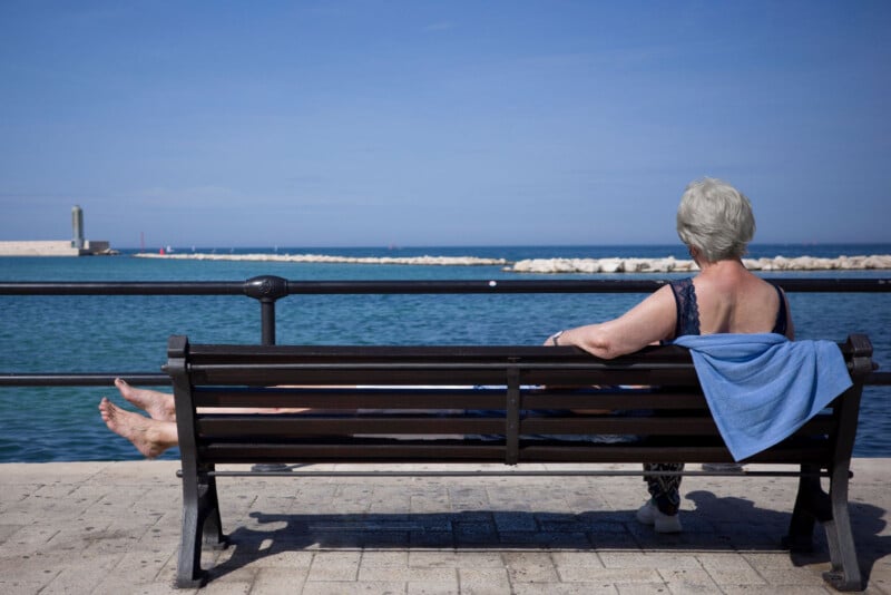 An older woman with short gray hair sits on a bench by the sea, facing the water. She has a blue towel draped over the bench and is barefoot, relaxing under a clear blue sky on a paved promenade.