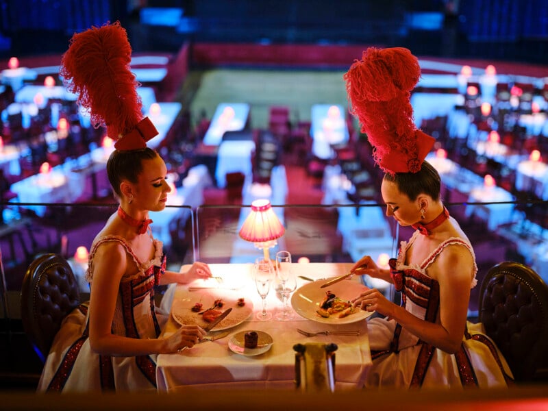 Two women in elaborate red feathered headdresses and costumes sit at a small restaurant table, eating dinner in a dimly lit, elegant venue with red lamps and tables set for an event in the background.