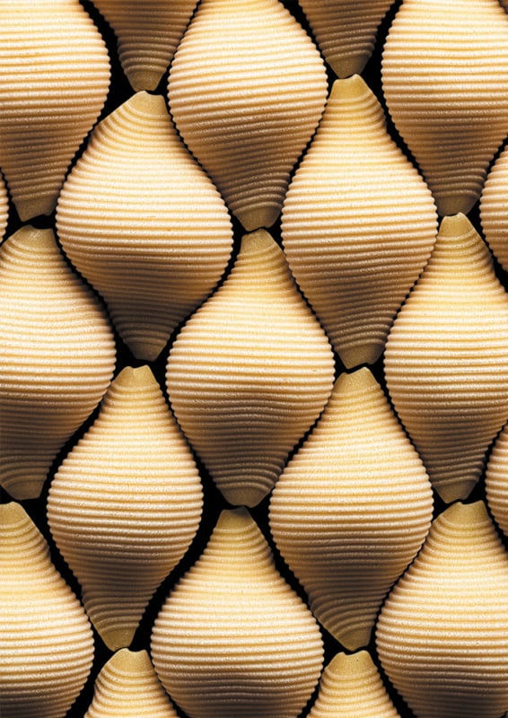 A close-up view of repeating, beehive-shaped objects with ridged, horizontal lines in warm shades of tan and gold, arranged in a dense, symmetrical pattern.